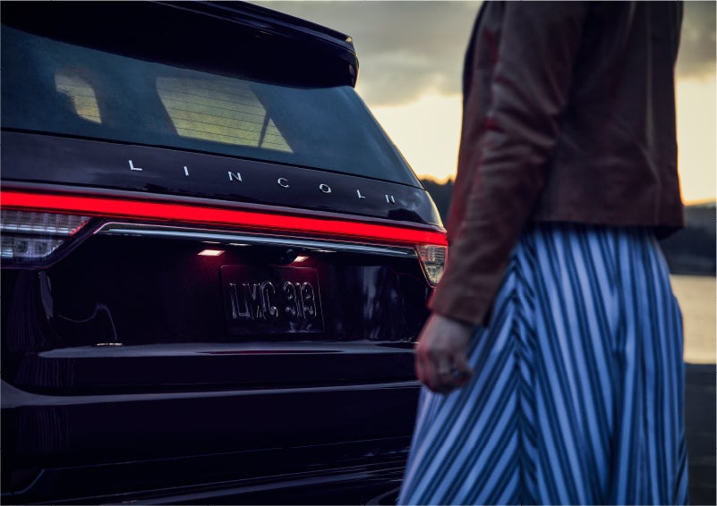 A person is shown near the rear of a 2023 Lincoln Aviator® SUV as the Lincoln Embrace illuminates the rear lights | McKie Lincoln, Inc. in Rapid City SD